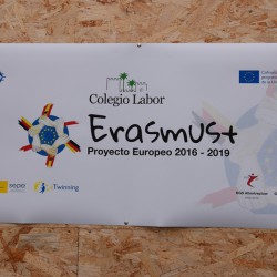 Zdjęcie przedstawia plakat Erasmus plus, który jest przyczepiony srebrnymi spinaczami do tablicy korkowej. Tło plakatu jest koloru białego. Napis Erasmus plus jest koloru czarnego. Logo stanowi koło koloru niebieskiego, na którym widać żółte gwiazdy oraz sześć trzymających się dłoni, które należą do ludzi z różnych krajów.