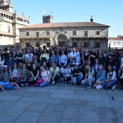 Zdjęcie grupy uczniów wraz z ich opiekunami. Znajdują się oni na placu w mieście Vigo w Hiszpanii. Jest słonecznie. Wszyscy są zadowoleni.
