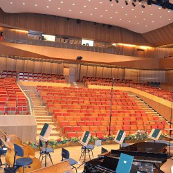 Obrazek przedstawia scenę wraz z miejscami siedzącymi w filharmonii. Krzesła są w kolorach czerwonym i pomarańczowym.