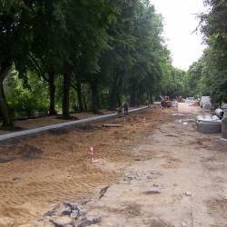 Budowa drogi rowerowej na ulicy Piastowskiej. Wokoło remontowanej drogi są rozłożyste drzewa. 