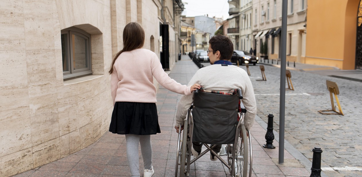 Na zdjęciu idzie dziewczynka i prowadzi wózek inwalidzki z osobą niepełnosprawną