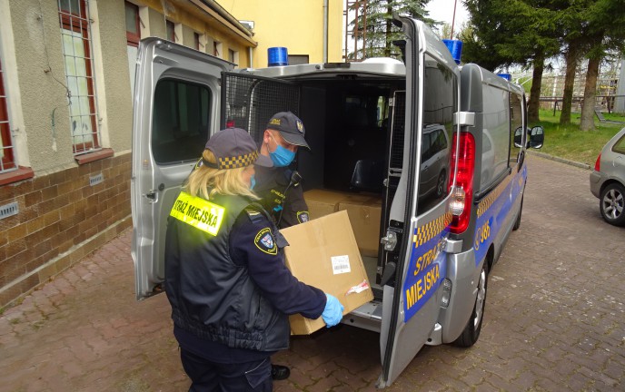 Dwoje Strażników Straży Miejskiej pakuje kartony z maseczkami ochronnymi do służbowego auta