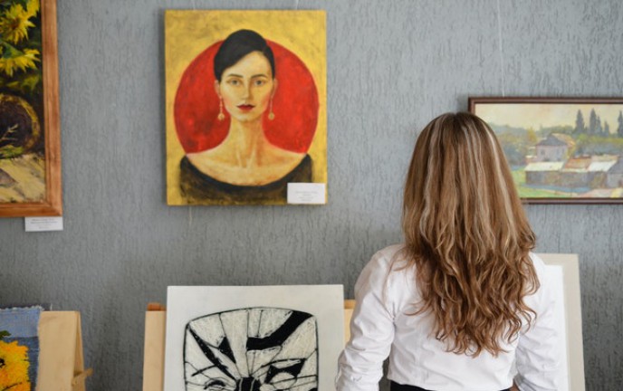 Zdjęcie przedstawia kobietę wpatrującą się w prace plastyczne, wiszące na ścianie, nieznanych artystów