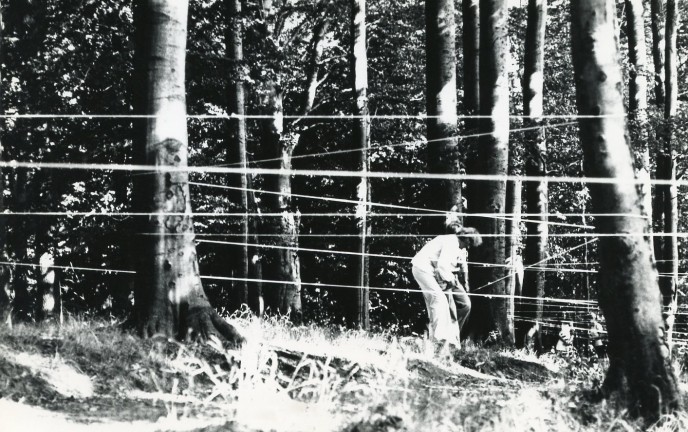 Czarno białe zdjęcie przedstawia mężczyznę w leśnej scenerii, który oplata sznurkiem widoczne na zdjęciu pnie drzew.