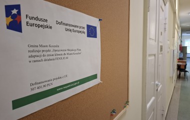 obrazem przedstawia w kolorze fragment korytarza urzędu oraz na pierwszym planie tablicę korkowa z plakatem informacyjnym dotyczącym projektu Opracowanie Miejskiego Planu Adaptacji do zmian klimatu dla Miasta Koszalina