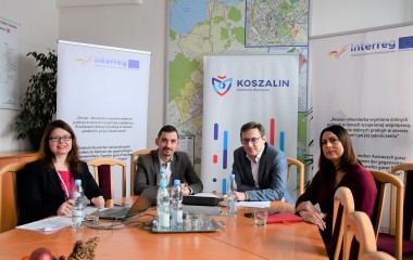 obrazek w kolorze przedstawia troje pracowników Urzędu Miejskiego w Koszalinie a także Sekretarza Miasta przy stole konferencyjnym, w tle mapy i banery na temat projektu