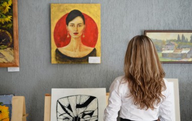Zdjęcie przedstawia kobietę wpatrującą się w prace plastyczne, wiszące na ścianie, nieznanych artystów