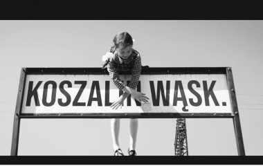 Wpadła bomba do piwnicy Grafika przedstawia czarno-biały kadr z teledysku, na którym widzimy małą dziewczynkę i tablicę Koszalińskiej Kolei Wąskotorowej
