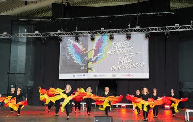 obrazek w kolorze przedstawia scenę na której tańczą kobiety wpomaranczowych spódnicach, nad sceną wisi baner z napisem Taniec bez granic w jezyku polskim i niemieckim a także znaki organizatorów