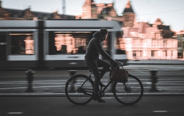 Obrazek ukazuje mężczyznę jadącego na rowerze, w tle tramwaj i zarysy budynków miejskich