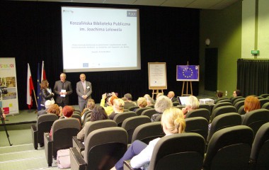 Obrazek w kolorze ukazuje grupę siedzących osób w auli biblioteki  ( widok z tyłu) w tle trzy osoby za nimi tablica z tytułem projektu ( slajd z prezentacji ) oraz flagi Polski, Niemiec i Unii Europejskiej 