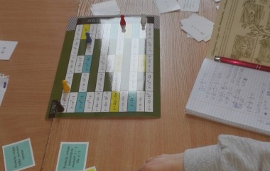 kolorowy obrazek ukazujący blat biurka szkolnego z rozłożonymi na nim materiałami dydaktycznymi : karteczkami, tabliczkami, zeszytami oraz dłonie dzieci uczących się za pomocą tych materiałów ( zajęcia w ZS1)